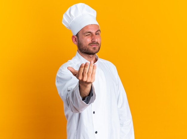 Confident young caucasian male cook in chef uniform and cap looking at camera winking doing come here gesture isolated on orange wall with copy space