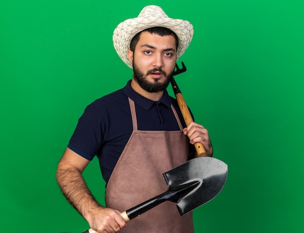 confident young caucasian male gardener wearing gardening hat holding rake and spade isolated on green wall with copy space