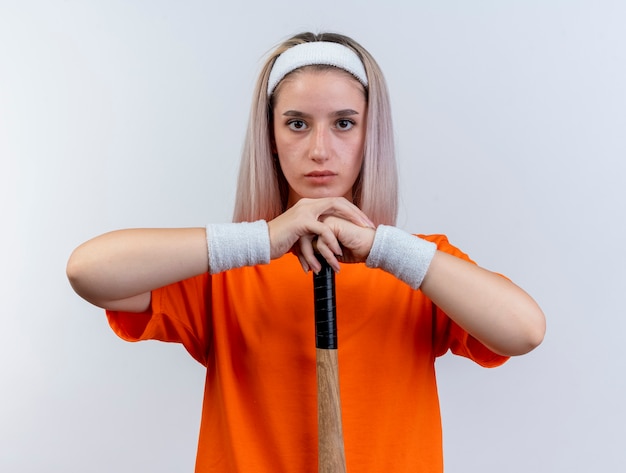 Confident young caucasian sporty girl with braces wearing headband and wristbands holds baseball bat upside down 