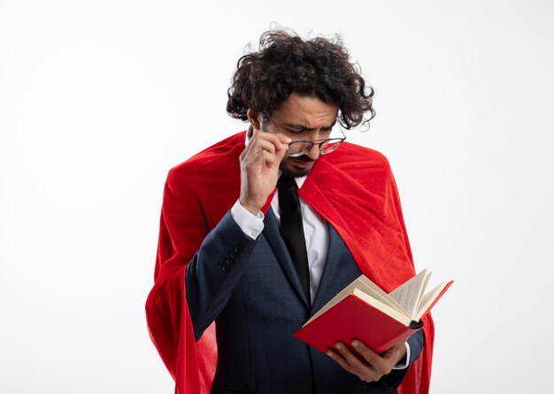 Confident young caucasian superhero man wearing suit with red cloak holds and looks at book through optical glasses 