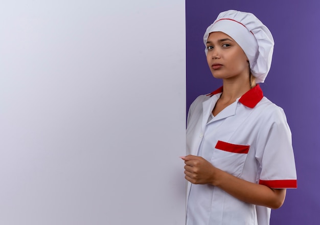 Confident young cook female wearing chef uniform holding white wall on isolated wall with copy space