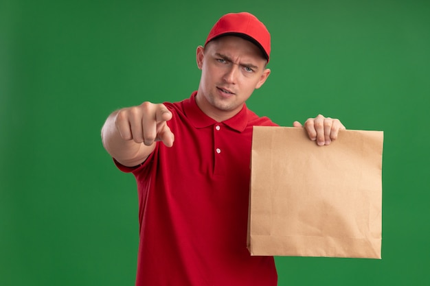 Confident young delivery man wearing uniform and cap holding paper food package showing you gesture isolated on green wall