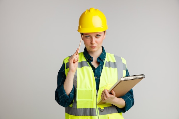 Confident young female construction worker wearing safety helmet and safety vest holding notepad doing think gesture with pencil 