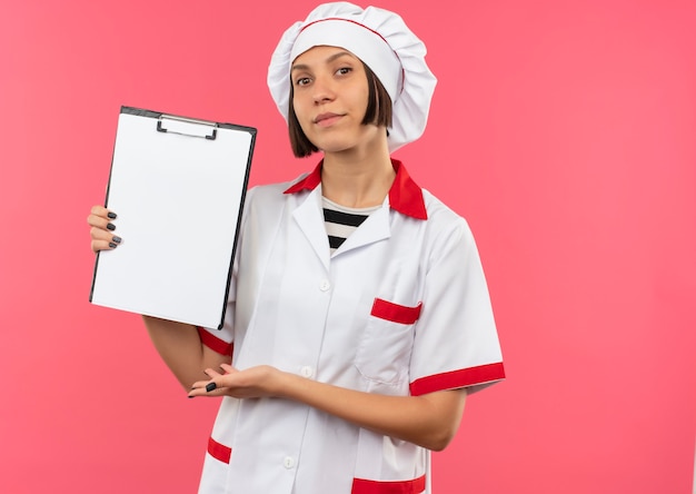 Free photo confident young female cook in chef uniform showing and pointing with hand at clipboard isolated on pink  with copy space