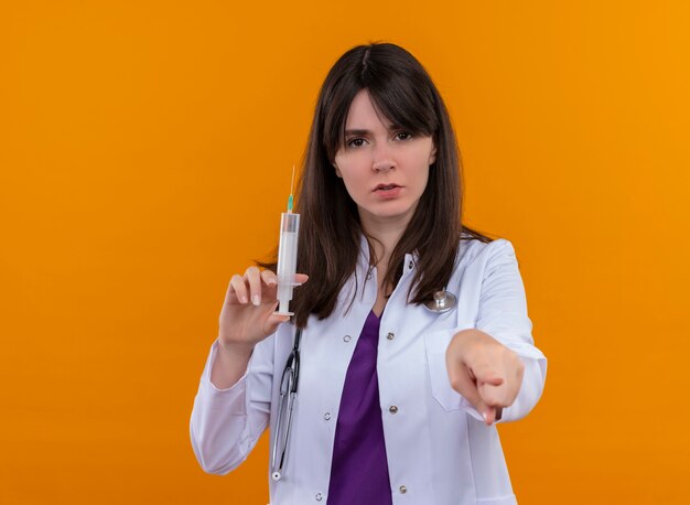 Confident young female doctor in medical robe with stethoscope holds syringe and points at camera on isolated orange background with copy space