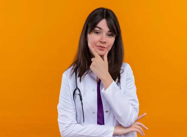 Confident young female doctor in medical robe with stethoscope puts hand on chin on isolated orange background with copy space