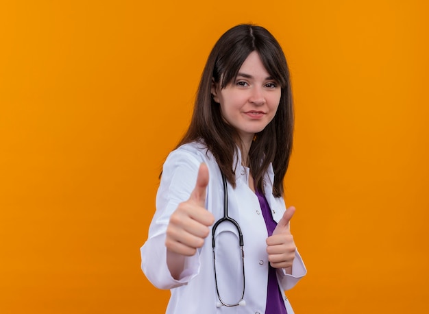 Confident young female doctor in medical robe with stethoscope thumbs up on isolated orange background with copy space