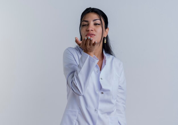 Confident young female doctor wearing medical robe looking sending blow kiss isolated