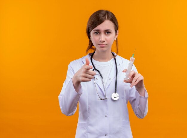 Confident young female doctor wearing medical robe and stethoscope holding syringe and doing you gesture on isolated orange space with copy space