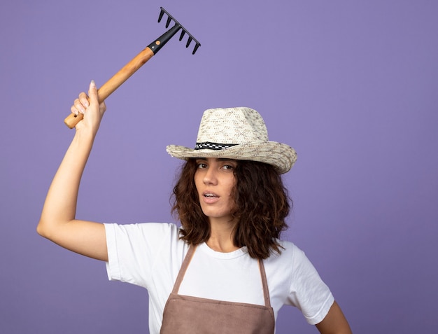 Free photo confident young female gardener in uniform wearing gardening hat raising rake