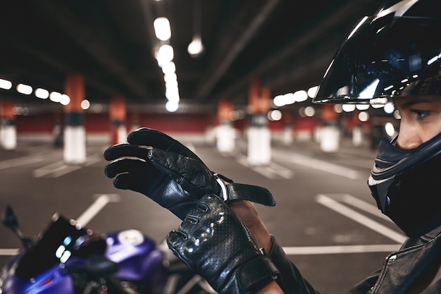 Free Photo confident young female racer wearing stylish motorcycle helmet putting on leather gloves, posing isolated in underground parking lot with her blue motorbike. selective focus on woman's hands