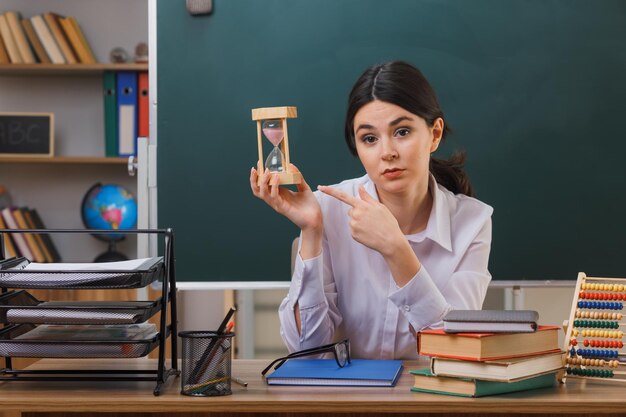 confident young female teacher holding and points at sand watch sitting at desk with school tools in classroom