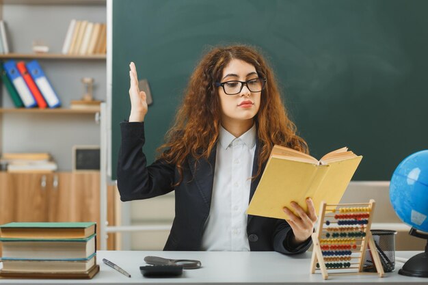 confident young female teacher wearing glasses holding and reading book sitting at desk with school tools in classroom