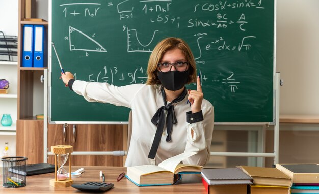 Confident young female teacher wearing glasses and medical mask sits at table with school tools points at blackboard with pointer stick in classroom