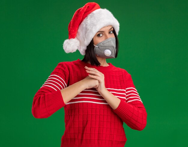 Free Photo confident young girl wearing santa hat and protective mask looking at camera doing greeting gesture isolated on green background