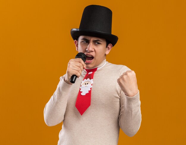 Confident young guy wearing hat with christmas tie speaks on microphone showing yes gesture isolated on yellow wall