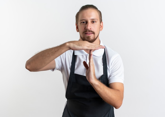 Confident young handsome barber wearing uniform doing timeout gesture isolated on white  with copy space