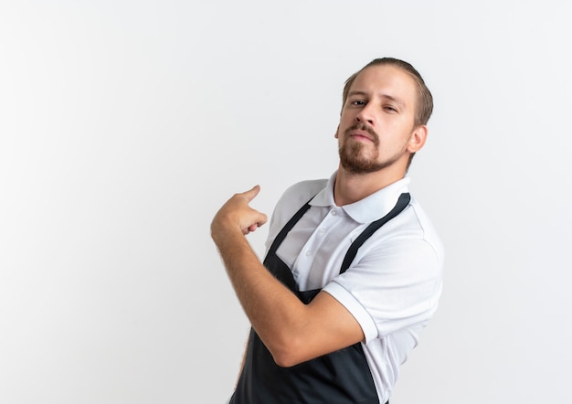 Confident young handsome barber wearing uniform pointing behind isolated on white  with copy space