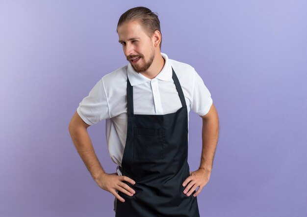 Confident young handsome barber wearing uniform winking and putting hands on waist isolated on purple  with copy space