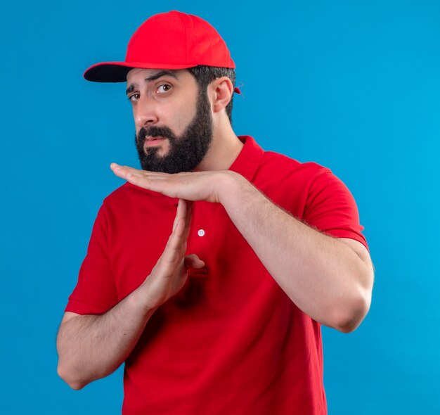 Confident young handsome caucasian delivery man wearing red uniform and cap doing timeout gesture isolated on blue 