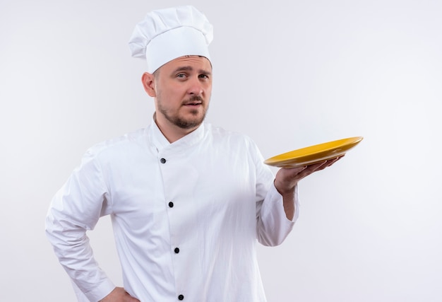 Free photo confident young handsome cook in chef uniform holding plate on isolated white wall