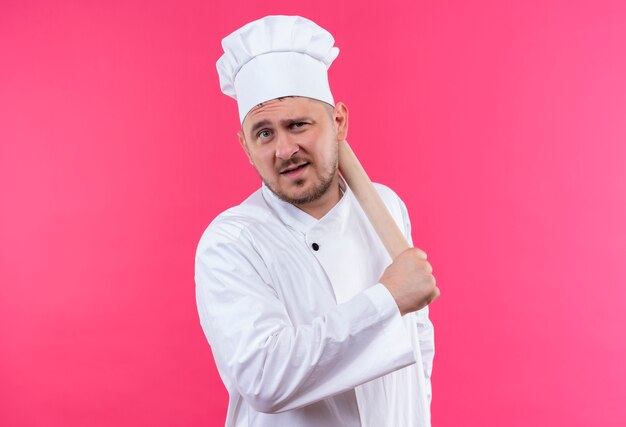 Confident young handsome cook in chef uniform holding rolling pin isolated on pink wall