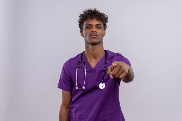 A confident young handsome dark-skinned doctor with curly hair wearing violet uniform with stethoscope pointing at camera with index finger 