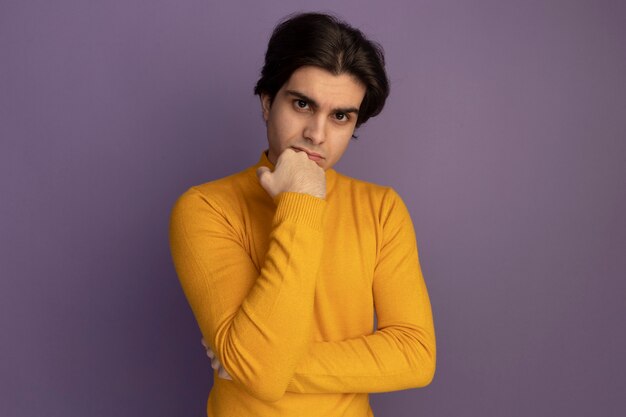 Confident young handsome guy wearing yellow turtleneck sweater putting fist on chin isolated on purple wall