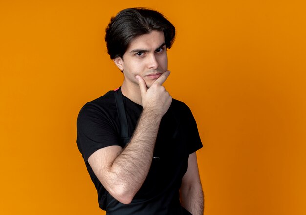 Confident young handsome male barber in uniform grabbed chin isolated on orange background