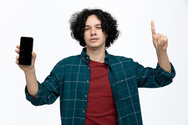 Confident young handsome man looking at camera showing mobile phone to camera raising index finger isolated on white background