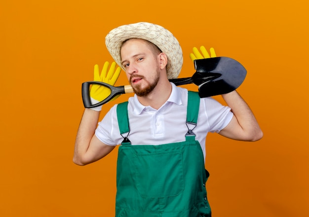 Confident young handsome slavic gardener in uniform wearing hat and gardening gloves holding spade behind neck isolated on orange wall with copy space