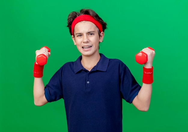 Confident young handsome sporty boy wearing headband and wristbands with dental braces holding dumbbells looking at front isolated on green wall