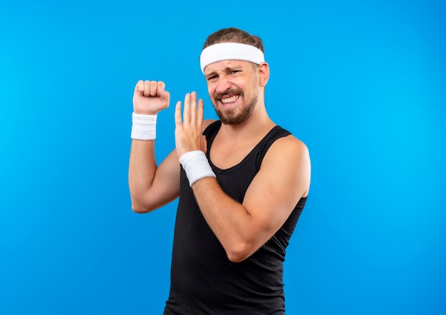 Confident young handsome sporty man wearing headband and wristbands clenching fist and pointing with hand at it isolated on blue wall with copy space