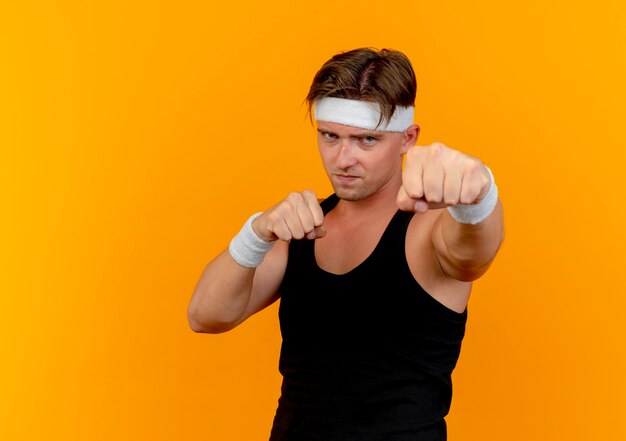 Confident young handsome sporty man wearing headband and wristbands doing boxing gesture isolated on orange  with copy space