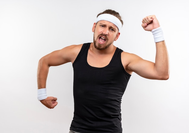 Confident young handsome sporty man wearing headband and wristbands gesturing strong looking  with open mouth isolated on white wall