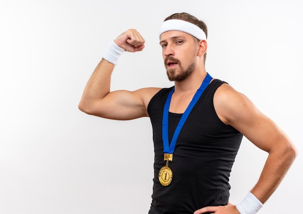 Confident young handsome sporty man wearing headband and wristbands gesturing strong with hand on waist isolated on white wall with copy space