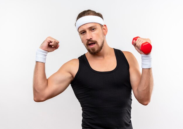 Confident young handsome sporty man wearing headband and wristbands holding dumbbell and gesturing strong isolated on white wall