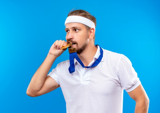 Confident young handsome sporty man wearing headband and wristbands and medal around neck holding and biting medal and looking at side isolated on blue wall