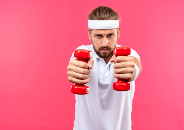 Free photo confident young handsome sporty man wearing headband and wristbands stretching out dumbbells towards  isolated on pink wall with copy space