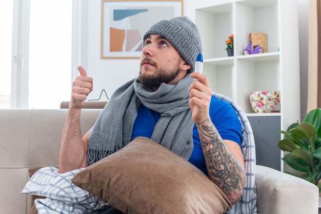 Confident young ill man wearing scarf and winter hat sitting on sofa in living room with pillow on his legs holding thermometer looking up showing thumb up