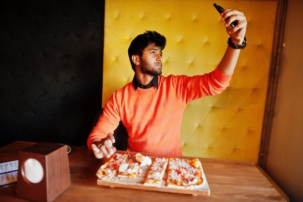 Free photo confident young indian man in orange sweater sitting at pizzeria eat pizza and makes photo on his phone