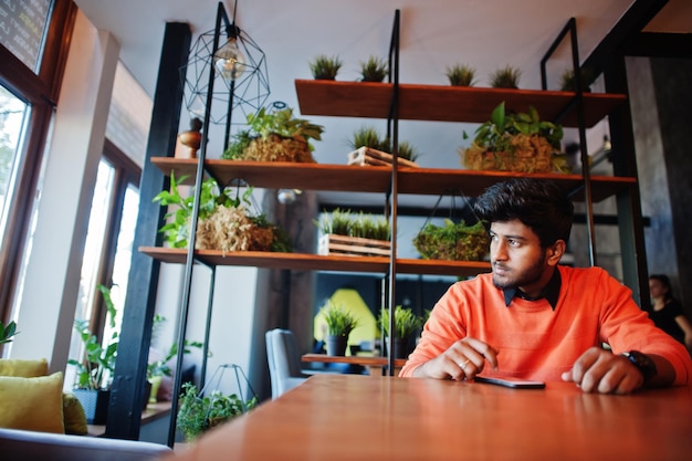 Confident young indian man in smart casual wear like orange sweater sitting at cafe and using his mobile phone