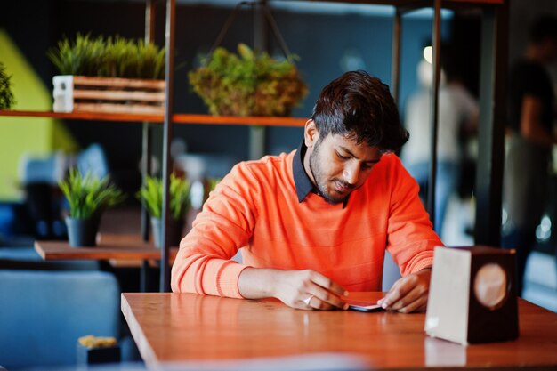 Confident young indian man in smart casual wear like orange sweater sitting at cafe and using his mobile phone