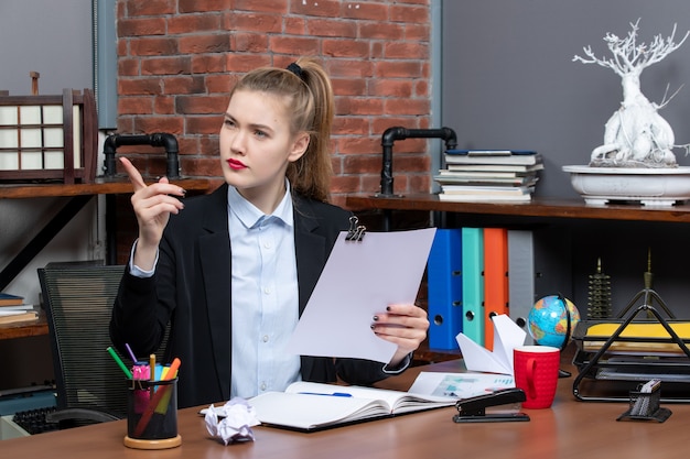 Confident young lady sitting at a table and reading her notes in notebook pointing up in the office