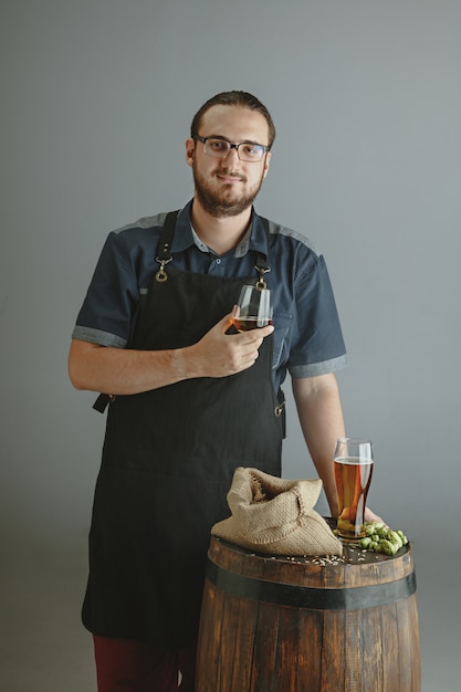 Free photo confident young male brewer with self crafted beer in glass on wooden barrel on grey background.