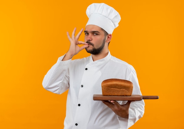 Confident young male cook in chef uniform holding cutting board with bread on it and doing tasty gesture isolated on orange wall