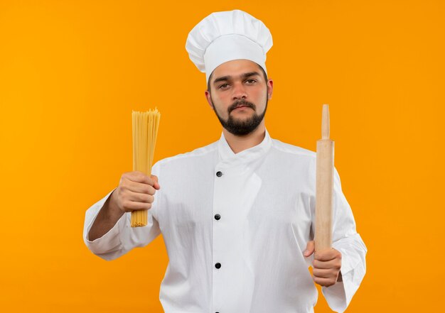 Confident young male cook in chef uniform holding spaghetti pasta and rolling pin isolated on orange wall