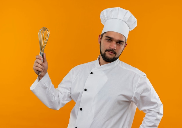 Confident young male cook in chef uniform holding whisk isolated on orange wall