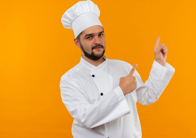 Confident young male cook in chef uniform pointing up isolated on orange wall with copy space