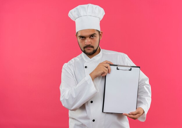 Confident young male cook in chef uniform showing clipboard isolated on pink wall with copy space